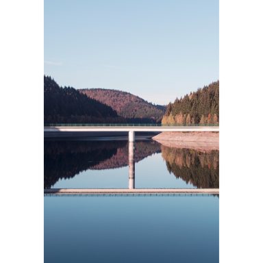 Landschaftsfoto der Talsperre Schönbrunn, im Mittelpunkt steht eine Brücke und ihre Spiegelung im Wasser, im Hintergrund sind Wald und Berge